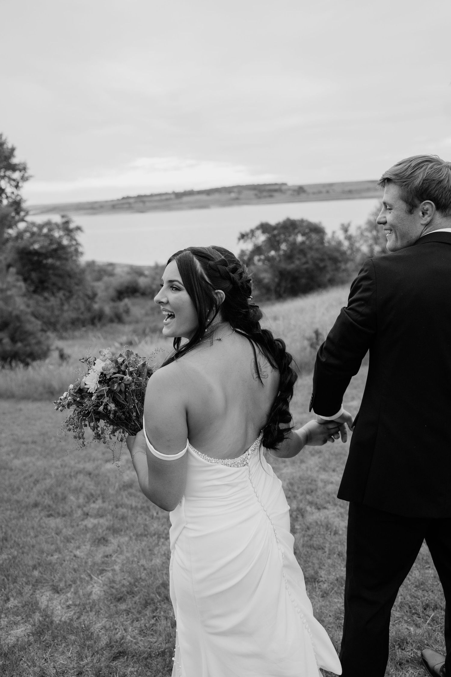 Black and white photo of laughing bride glancing back while holding grooms hand near a lake.