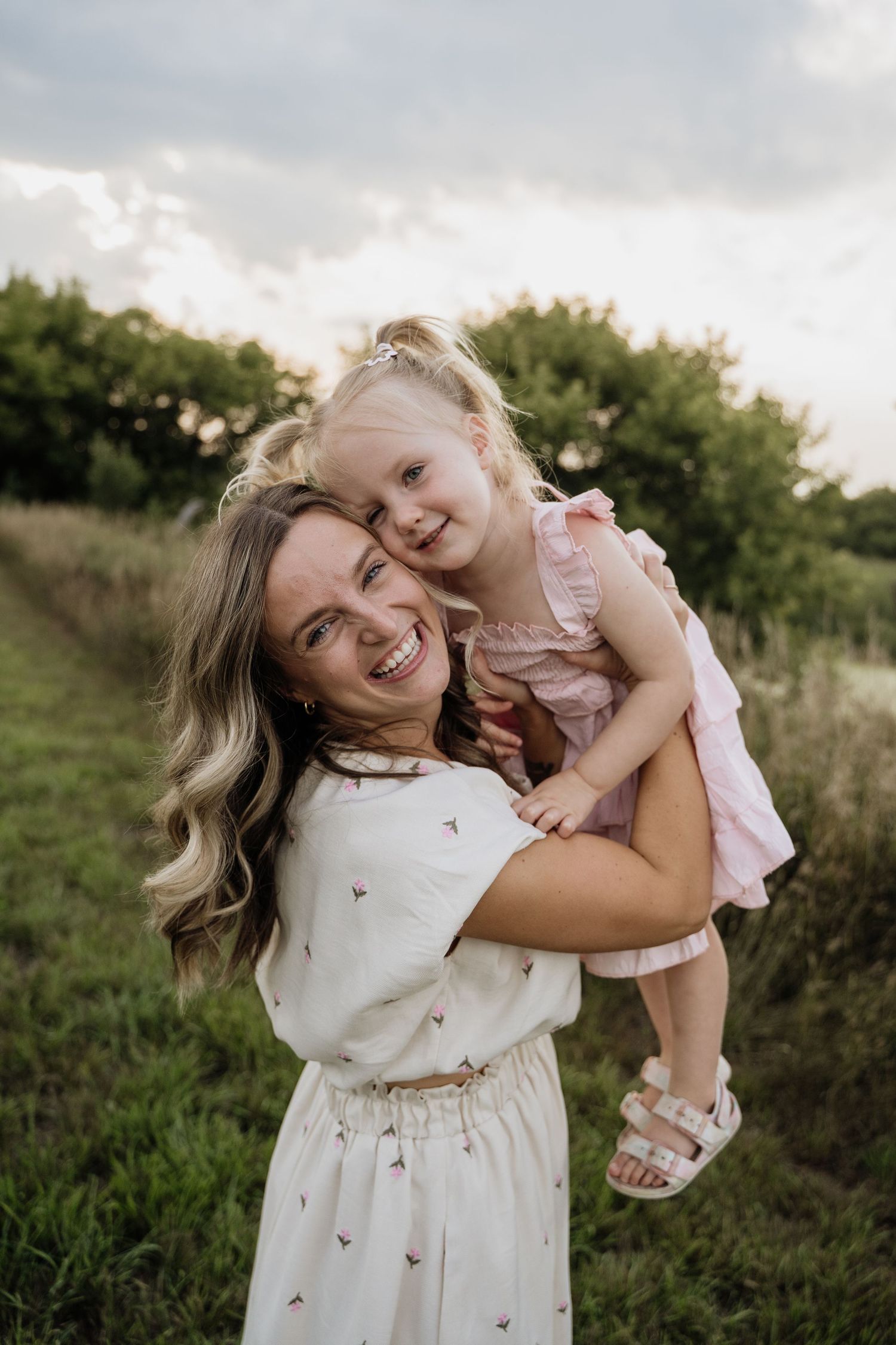 Laughing mother holding her smiling toddler daughter on her back in a green outdoor setting at dusk.