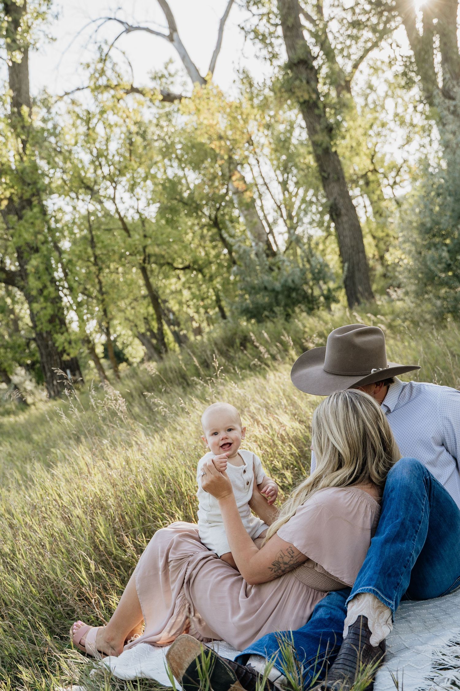 Young family sitting together in sunny green meadow, parents enjoying moment with smiling baby.