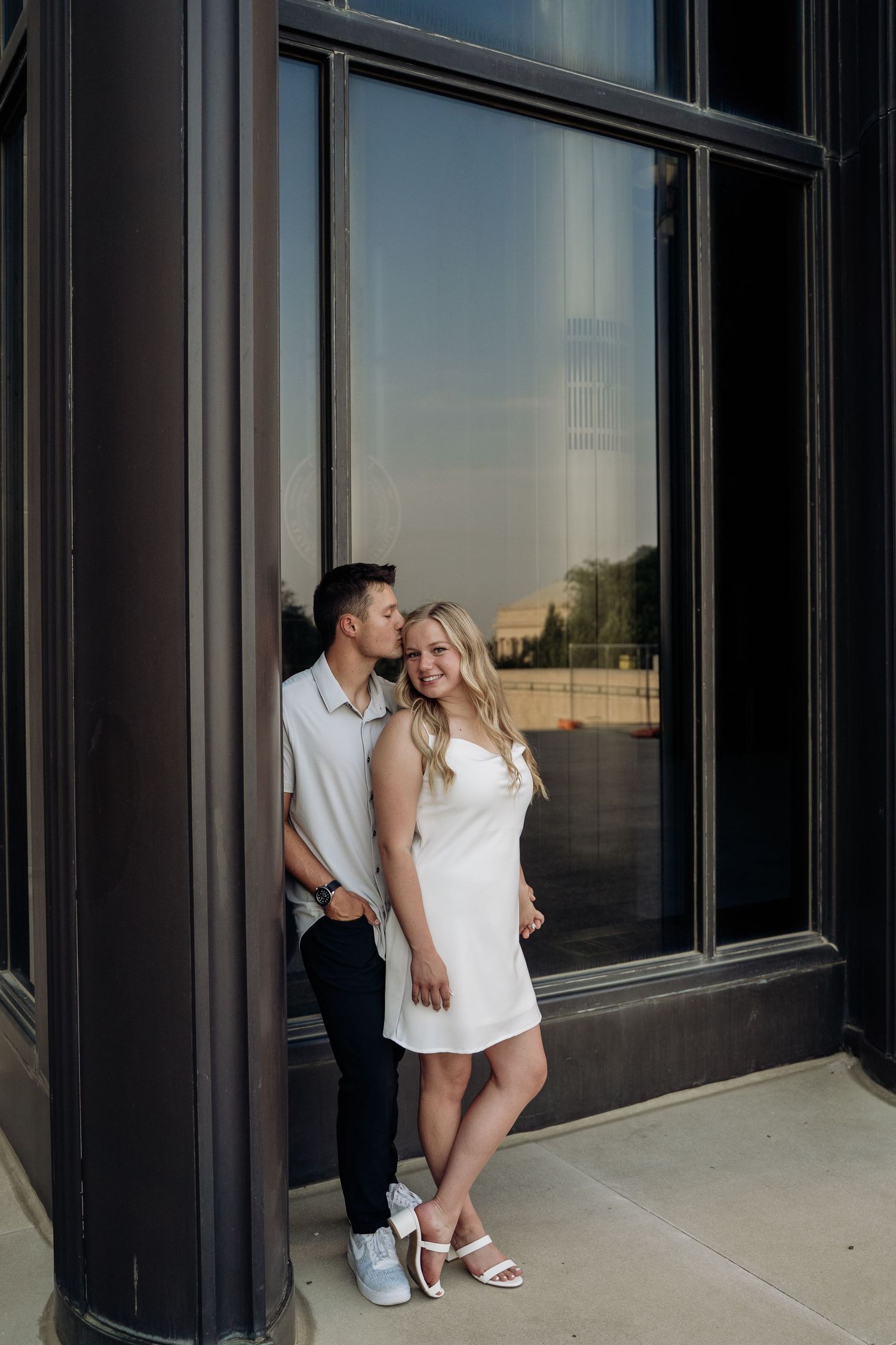 Couple posing by dark building window, man kissing womans cheek as she smiles at camera.