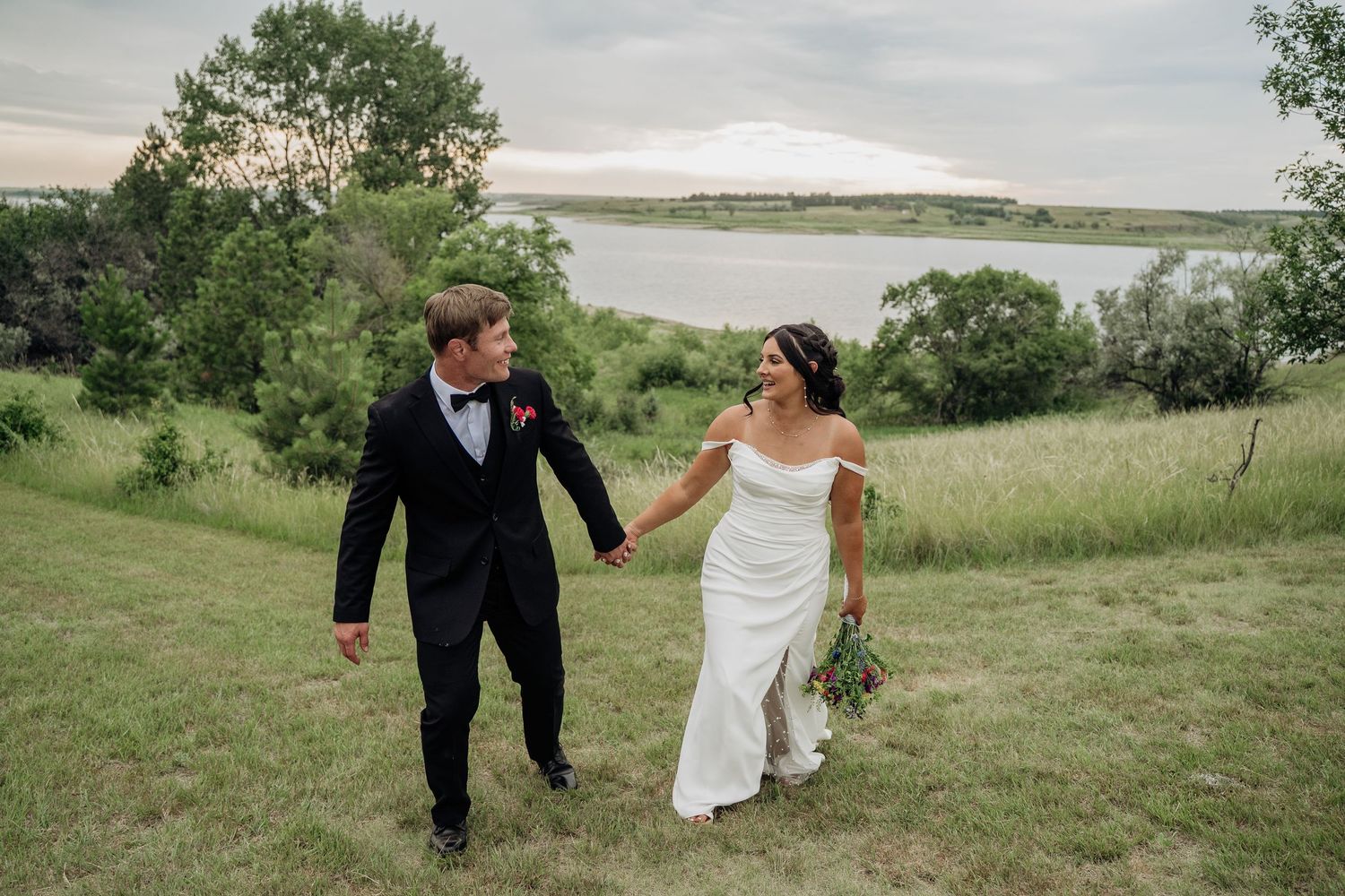 Bride and groom holding hands walking through green meadow with a beautiful lake backdrop.