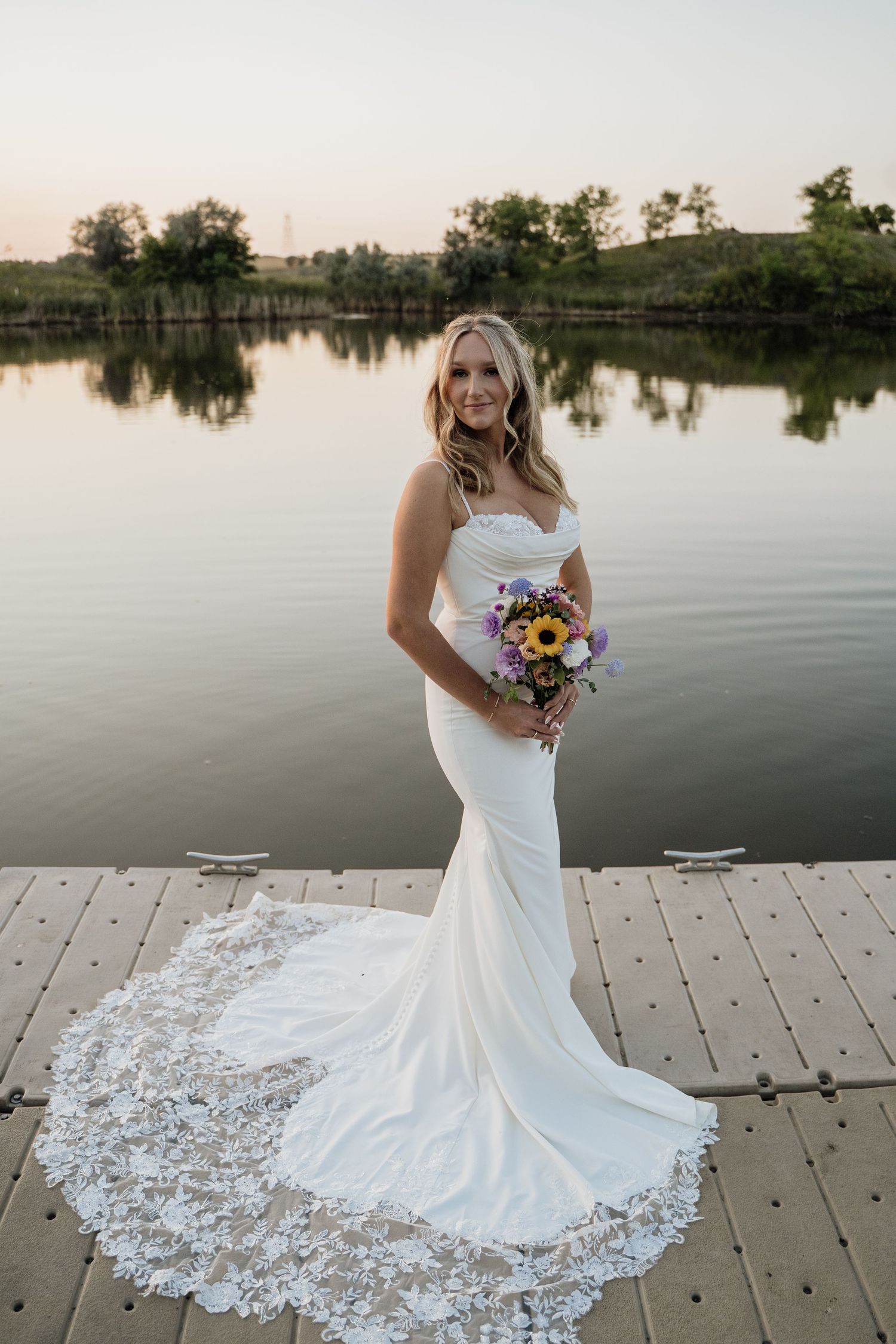 Bride in a mermaid gown with lace train holding a colorful bouquet on a dock at sunset.