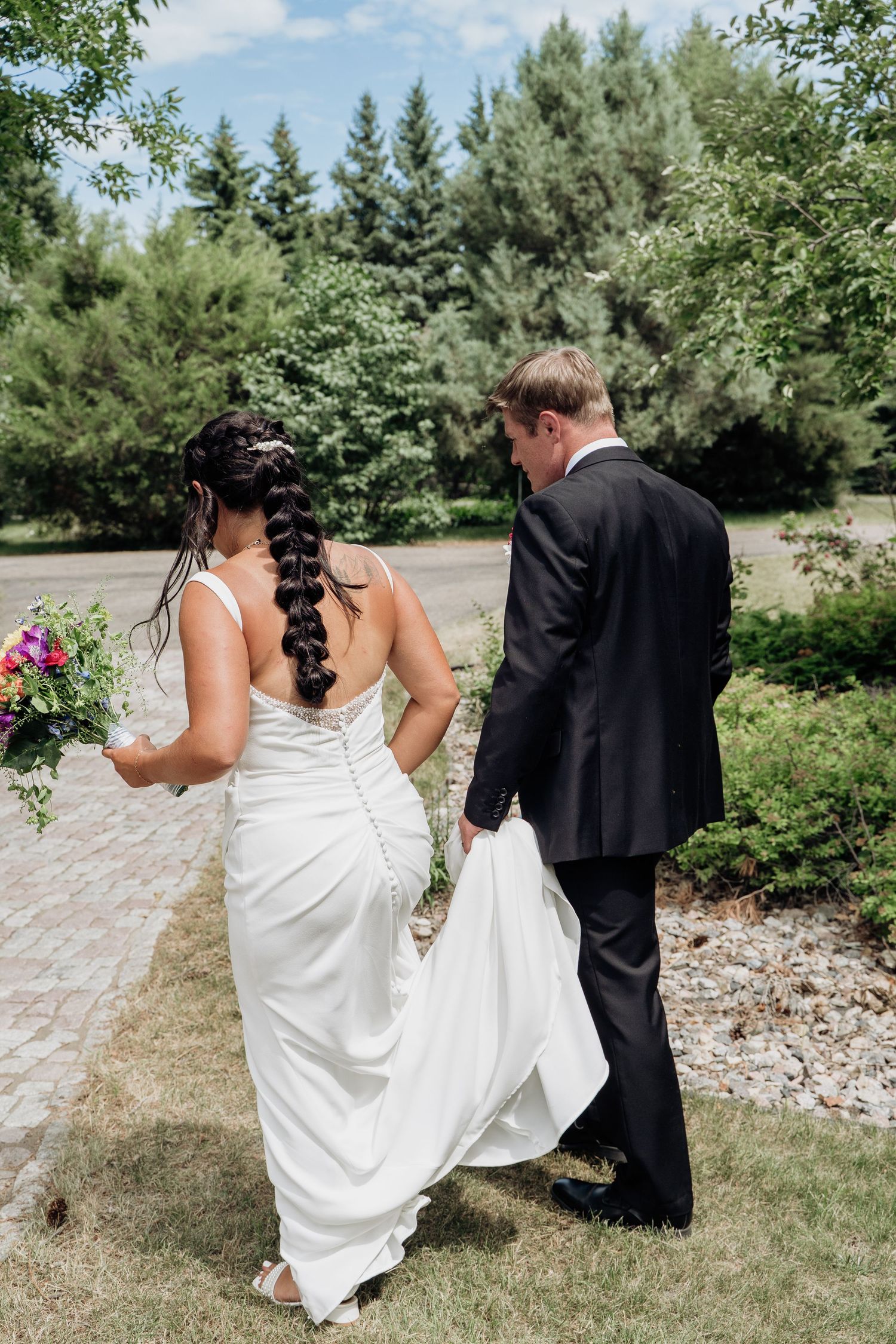 Bride and groom walking through garden, groom carrying wedding dress train.