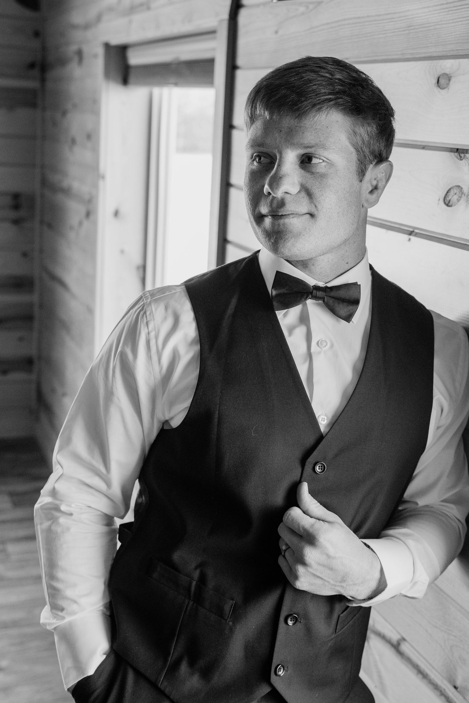 Black and white portrait of a groom in a vest and bow tie adjusting his jacket in a rustic wooden room.