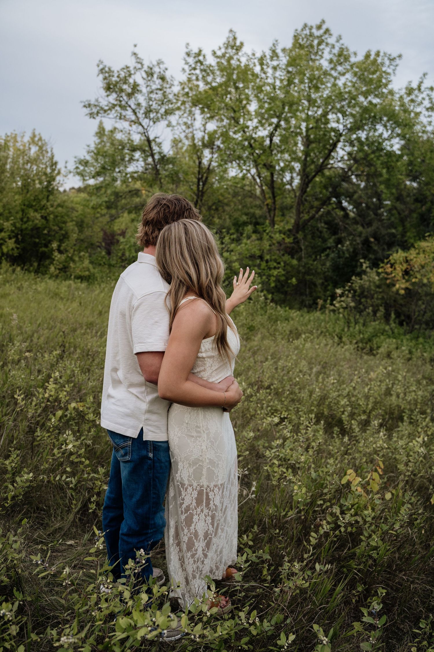 A couple embraces in a lush green field, the woman in a white lace dress showing her engagement ring.