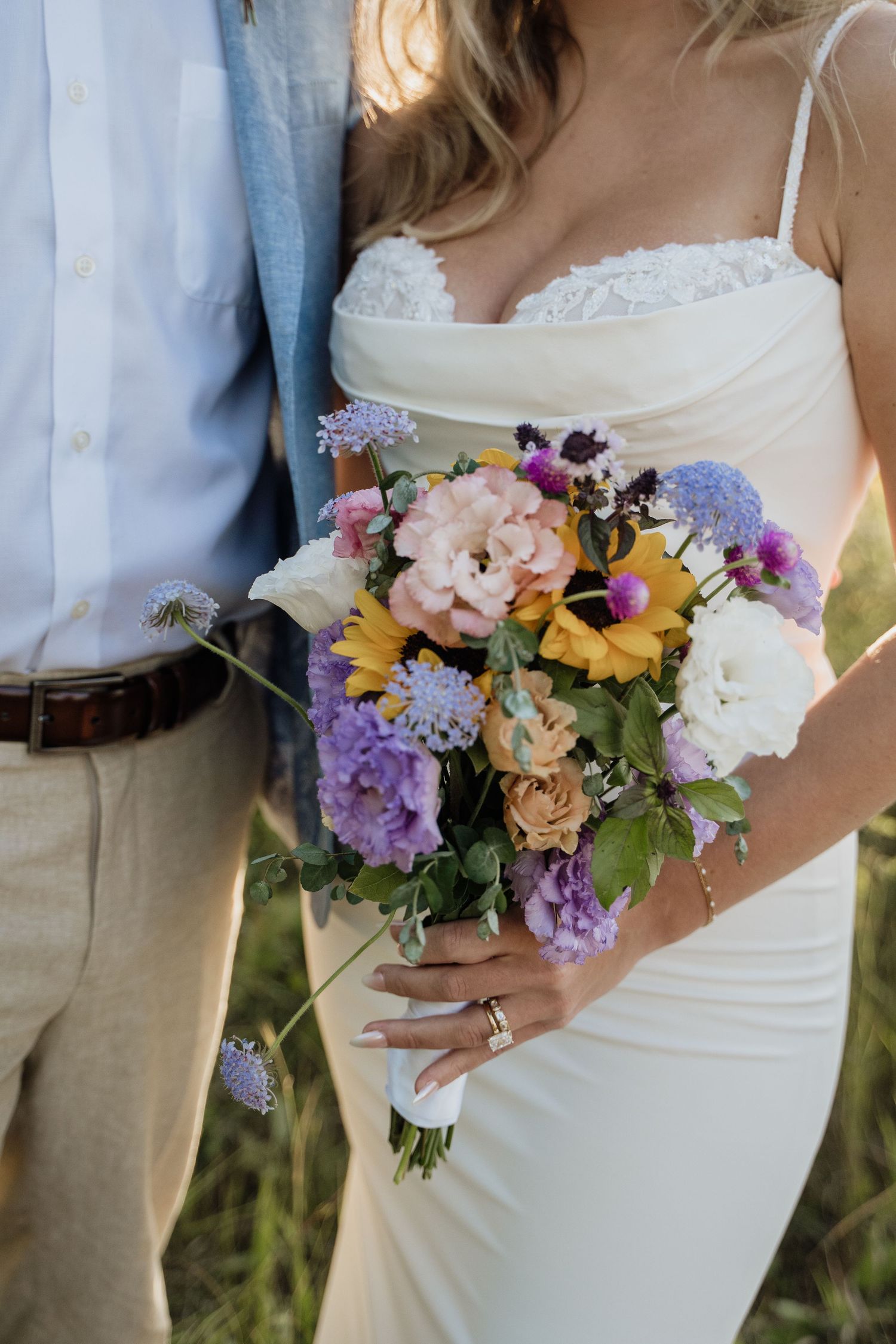 Bride holding a colorful wedding bouquet with sunflowers, lavender, and peonies in a white lace gown.