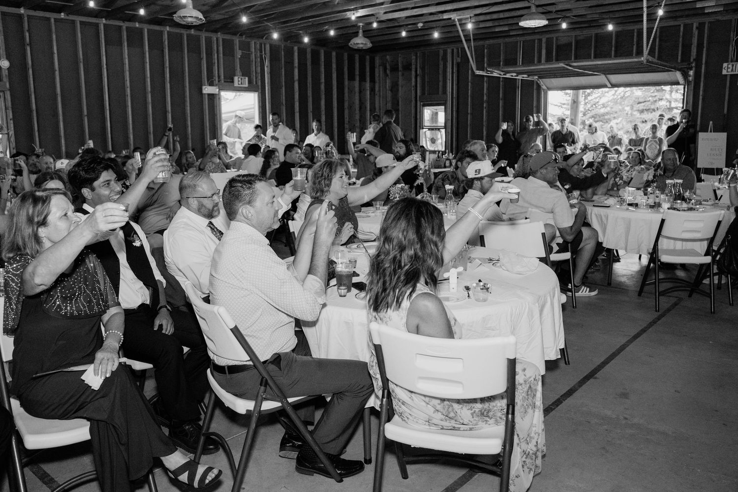 Black and white photo of wedding reception guests raising glasses in a toast inside a decorated barn venue.