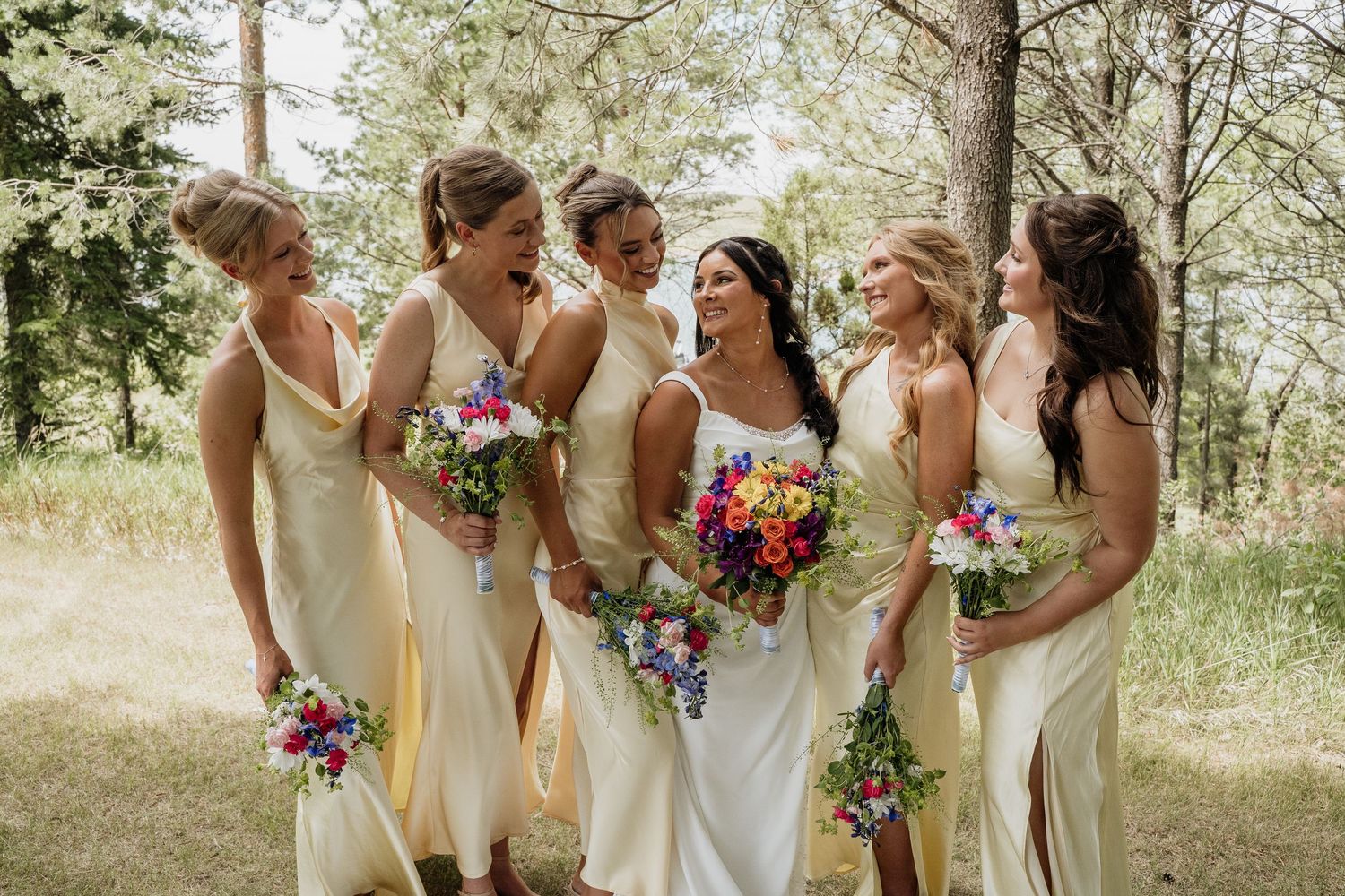 Bride and five bridesmaids in cream dresses laughing together outdoors in a wooded setting.