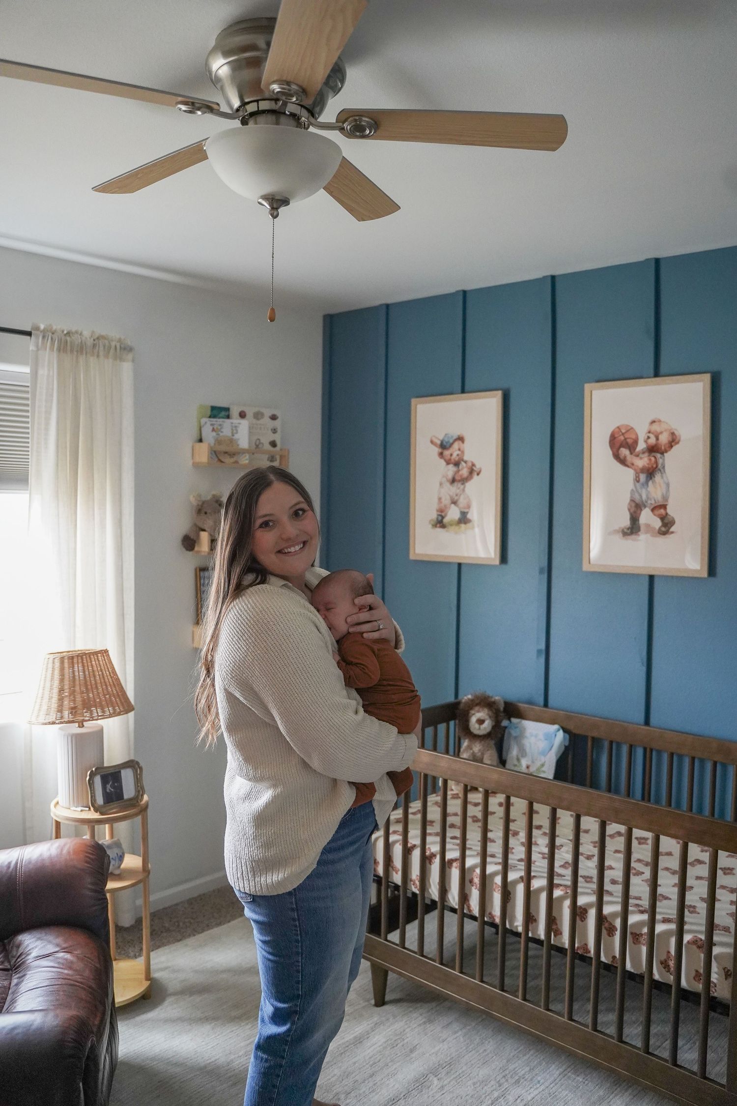 Mother holding newborn baby in a blue nursery with wooden crib and framed artwork.