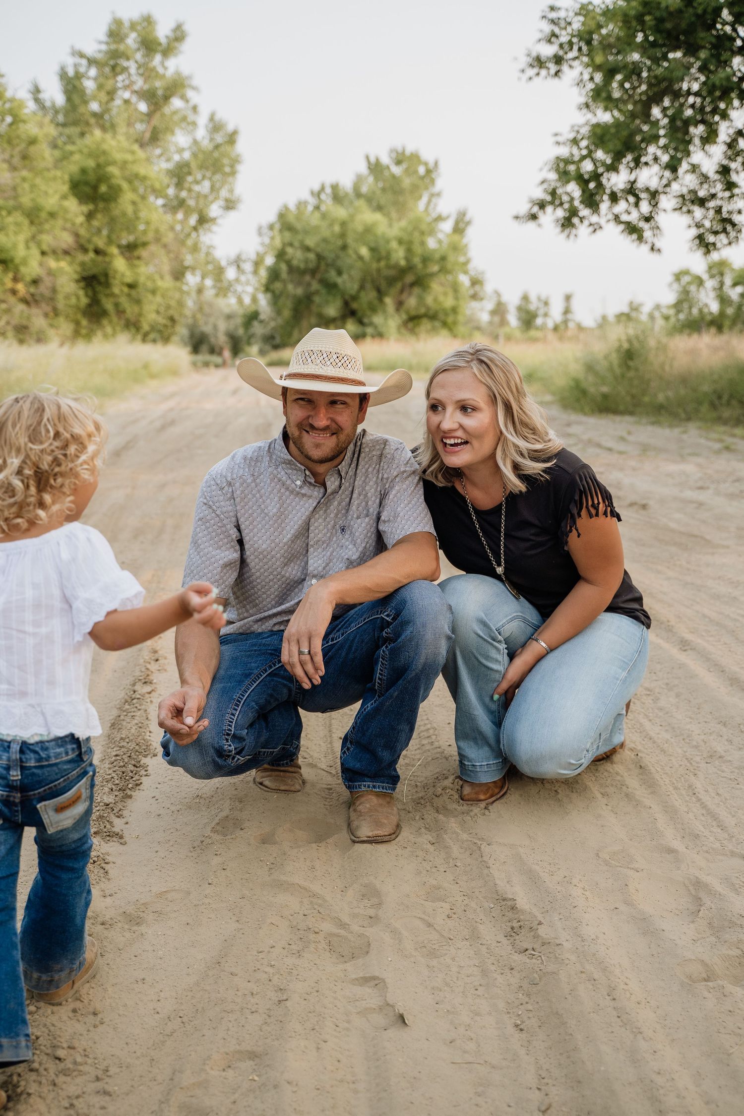 Parents in cowboy hats crouching on sandy path smiling at their young blonde toddler.