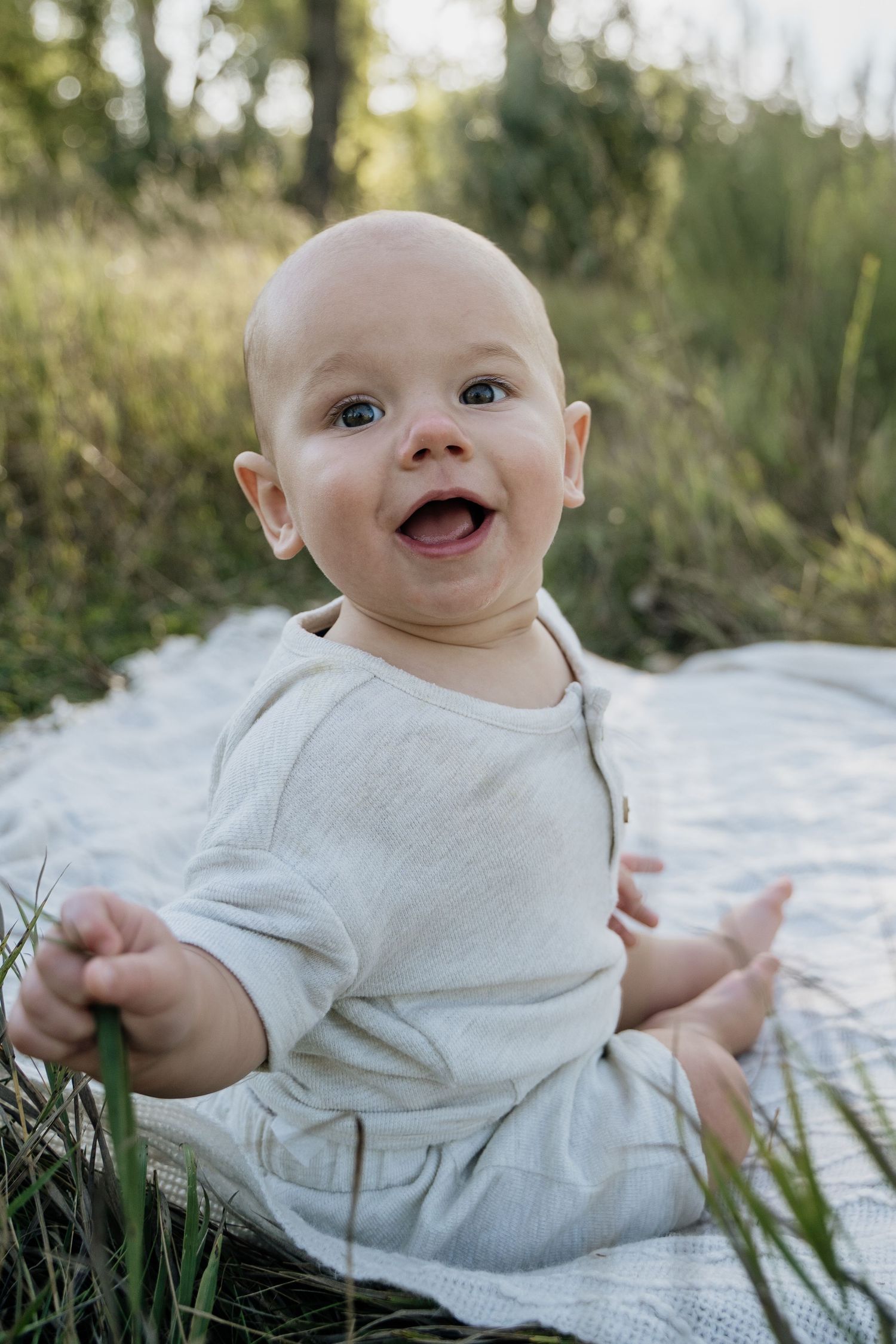 Adorable bald baby in white onesie sitting on blanket in green grass, smiling joyfully.