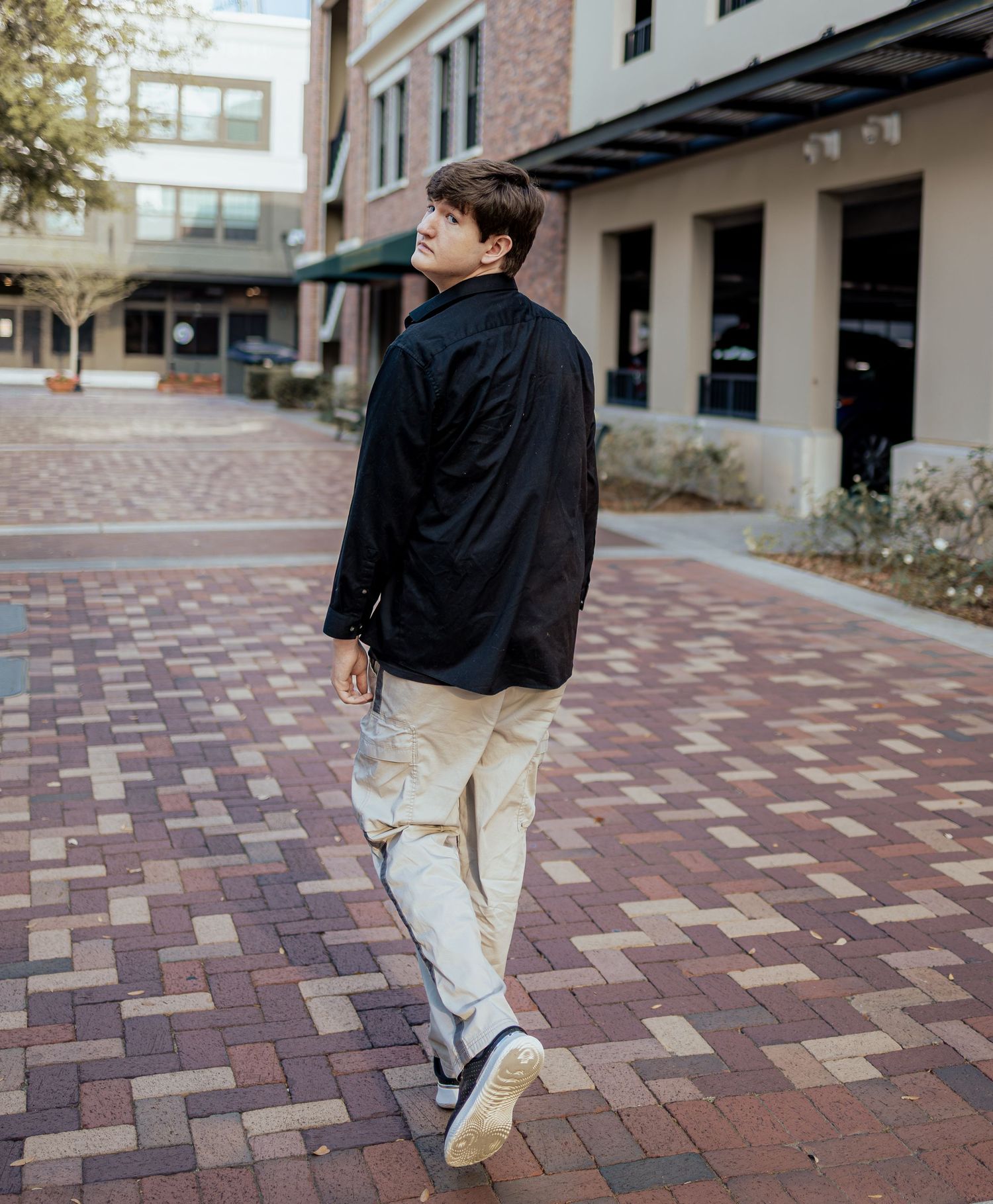 Person in black jacket and khaki pants walking on brick pavement near modern building exterior.