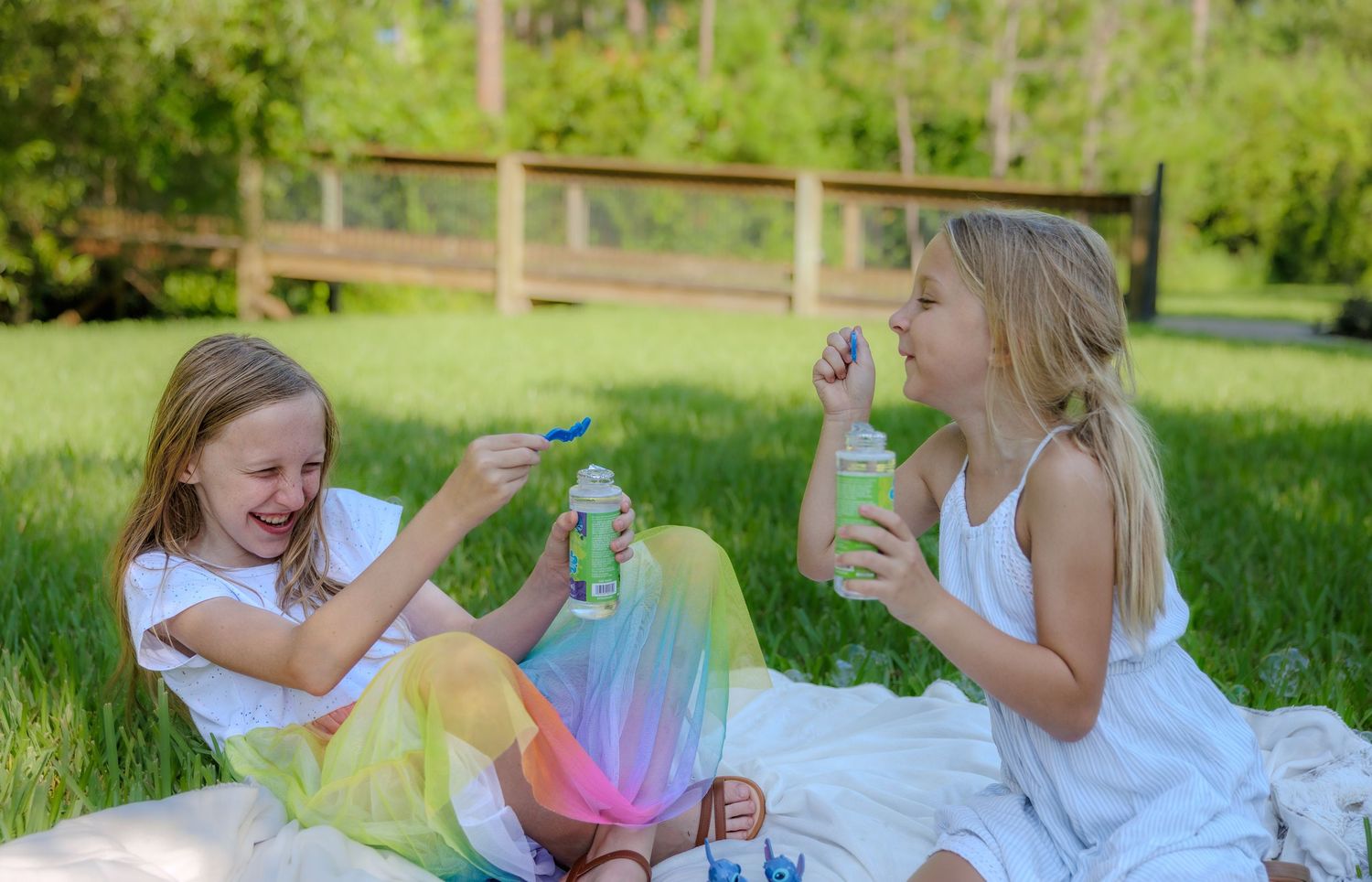 Two young girls in white dresses sit on a blanket in a sunny park, blowing bubbles and holding drink bottles beside a wooden fence.