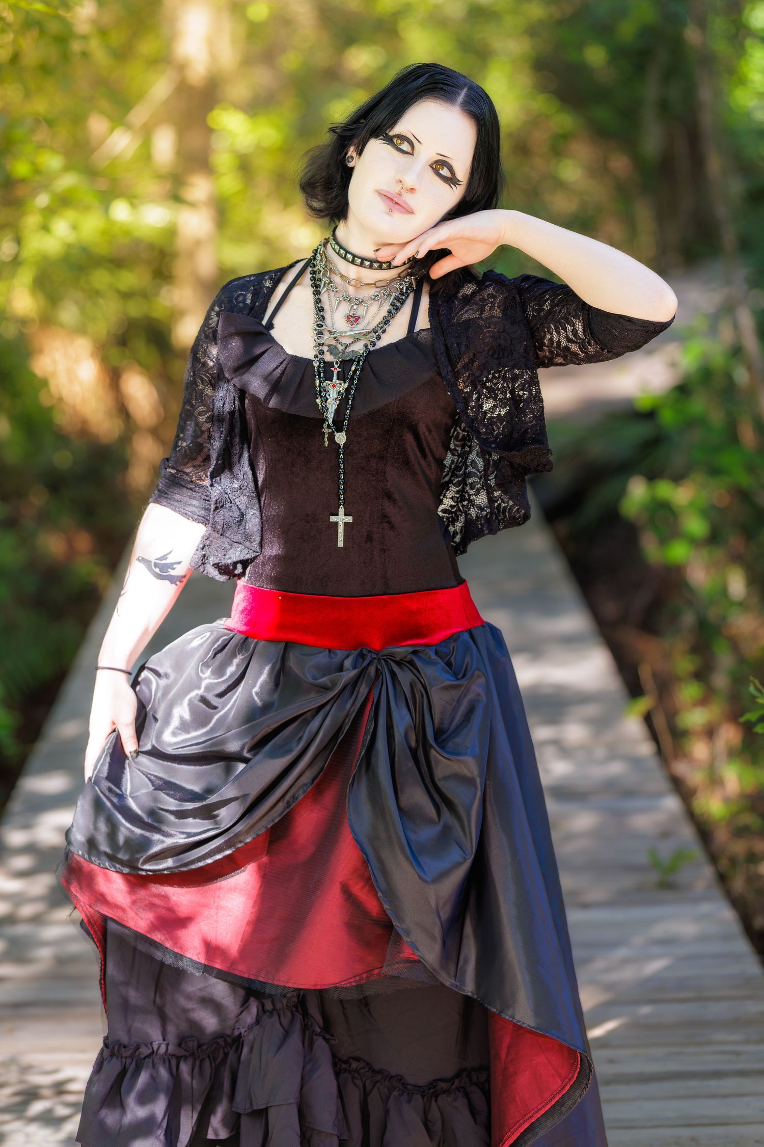 A gothic woman in black lace top, corset, and dramatic black skirt with red sash poses on a wooden bridge.
