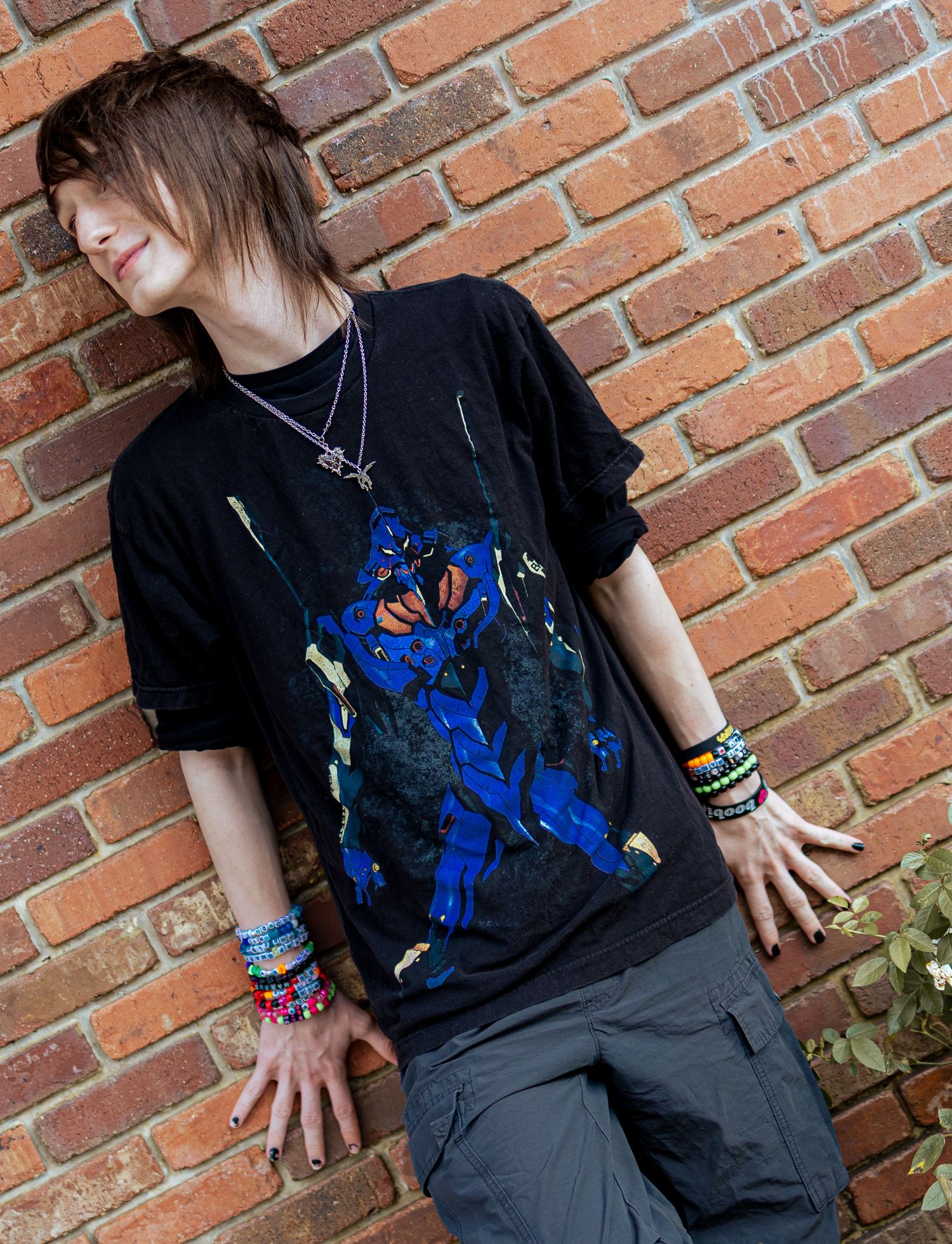 Person in black t-shirt with blue design poses against brick wall wearing dark pants and bracelets.