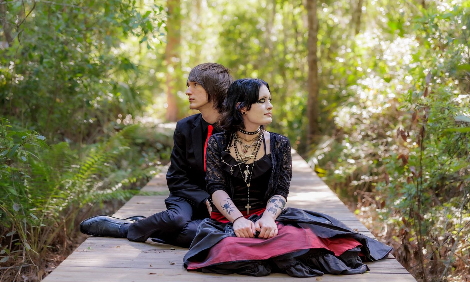 Two people in dark gothic clothing sit back to back on a wooden boardwalk surrounded by green forest foliage.