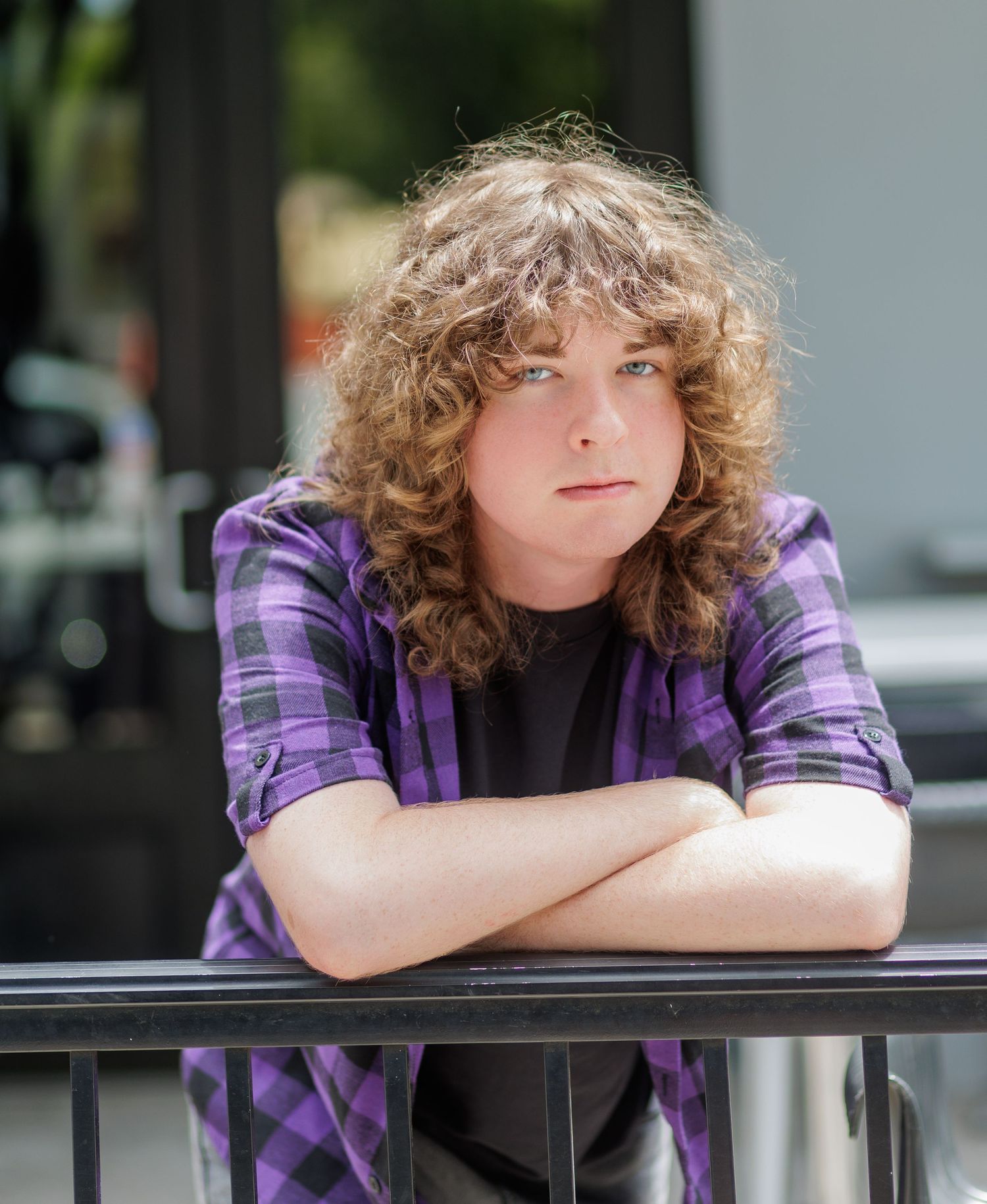Young person with long curly hair and a purple plaid shirt leans on a black railing with arms crossed, looking at the camera outdoors.