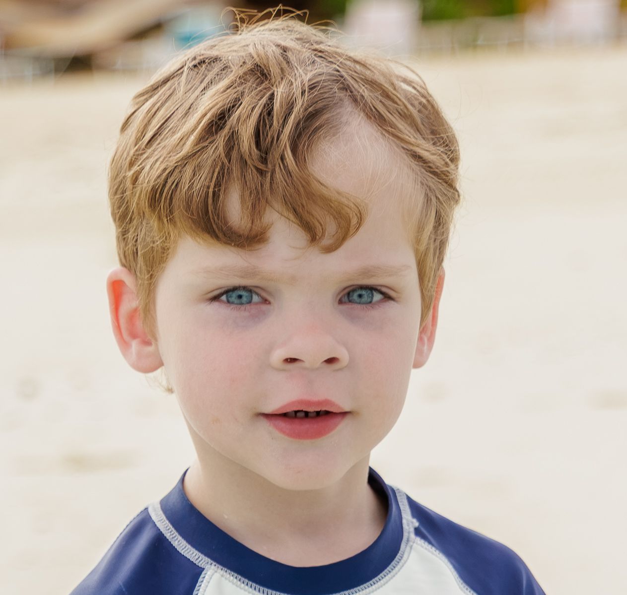 Close-up portrait of a young blond boy with blue eyes wearing a navy and white rash guard on a sandy beach.