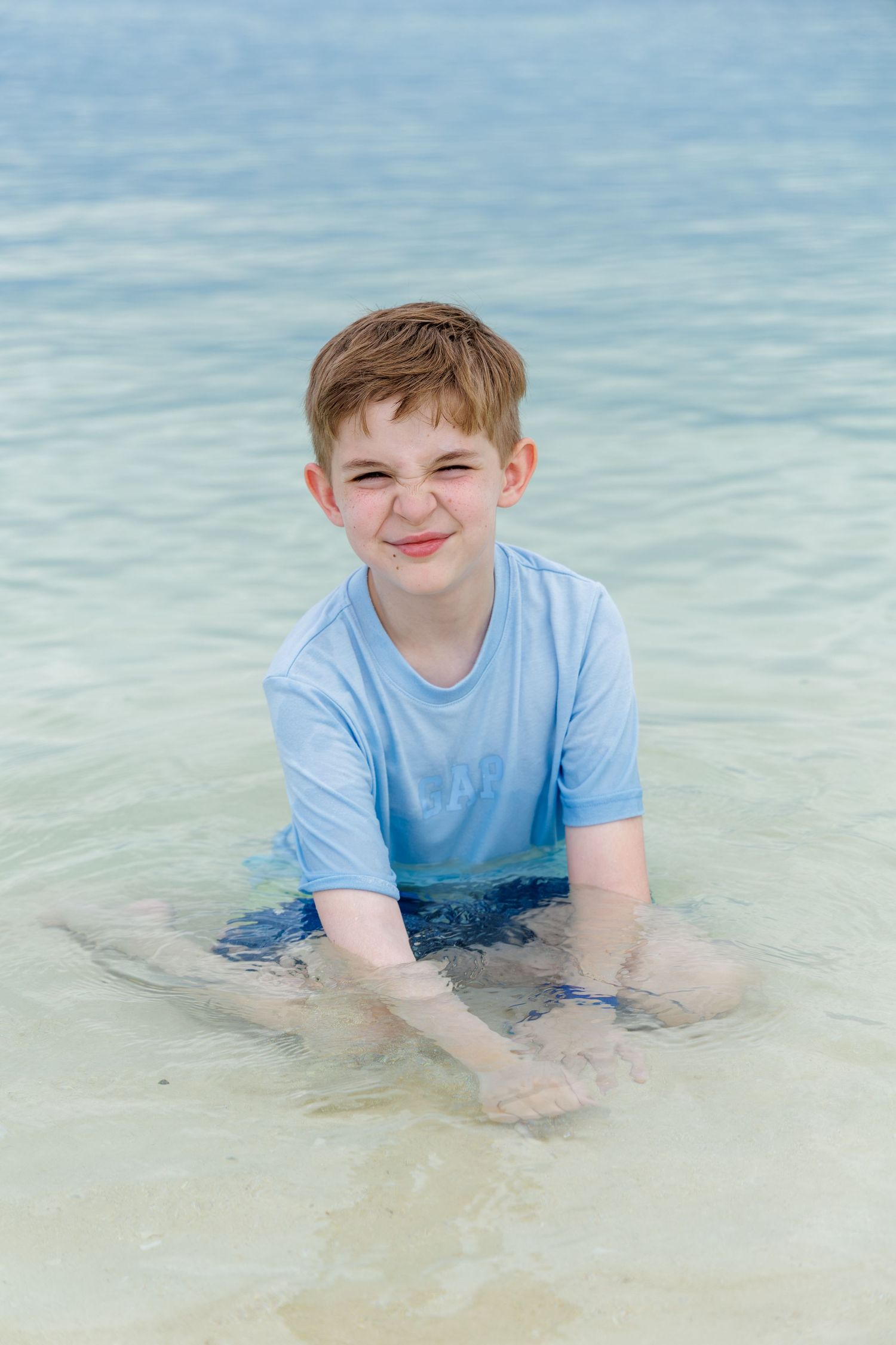 Smiling boy in a light blue rash guard squints playfully while sitting in shallow, clear turquoise water at a tropical beach.
