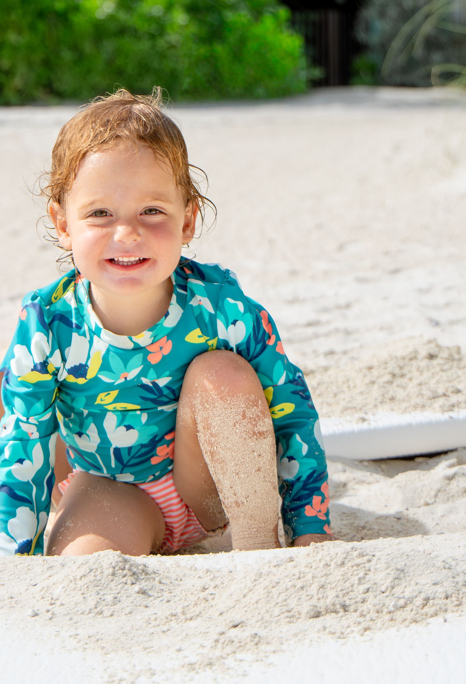 Child playing in white sand at the beach wearing a turquoise patterned swimsuit and creating sandcastles.
