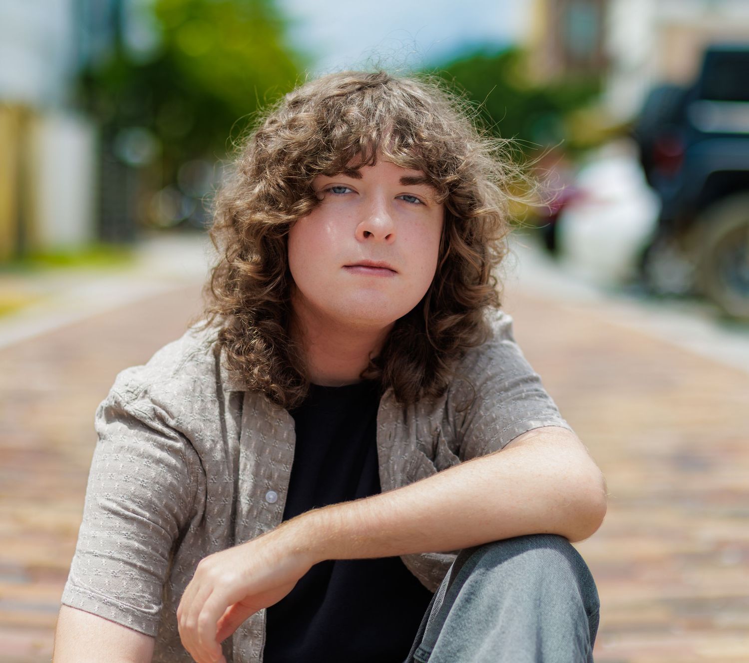 Young person with curly shoulder-length hair sits outdoors on a brick street, wearing a gray button-up shirt over a black tee.