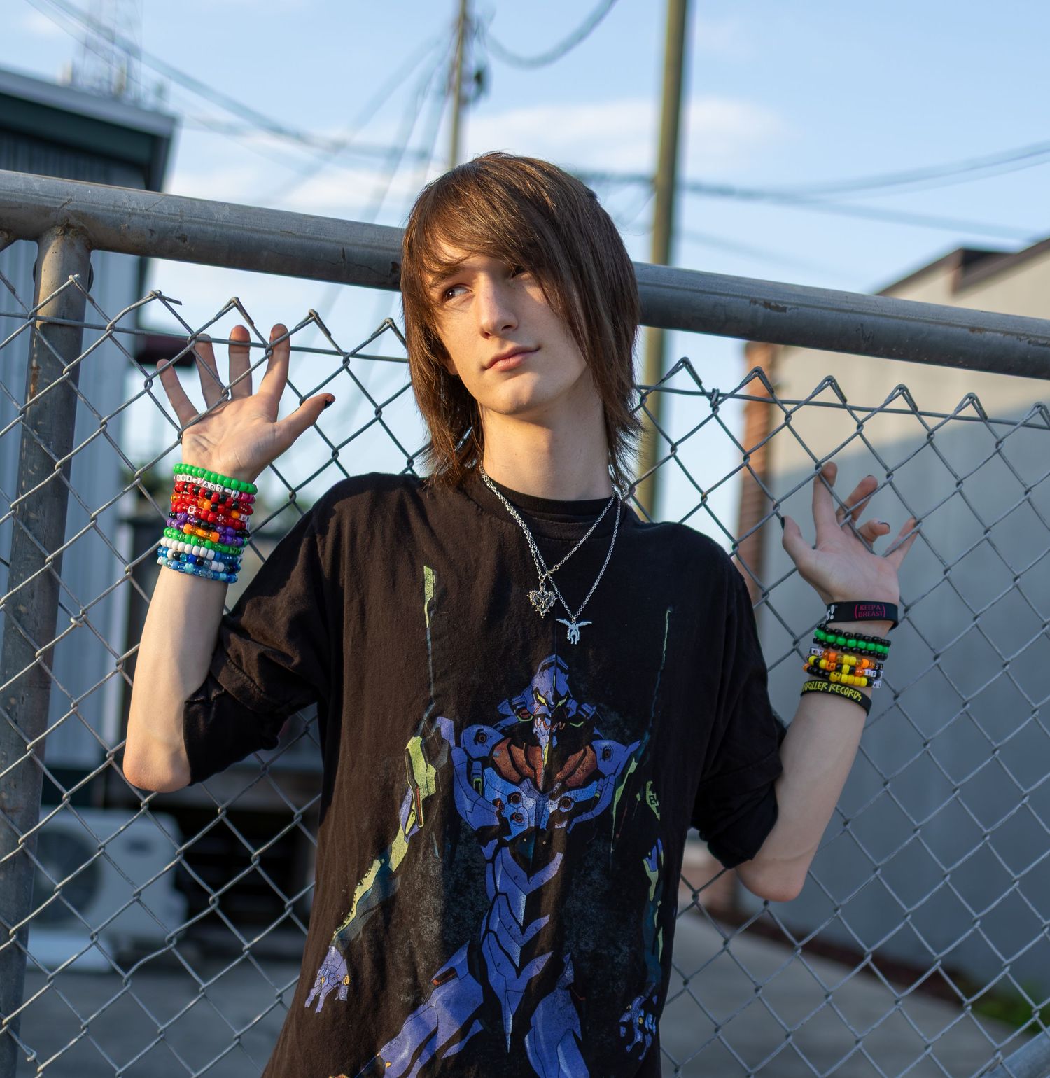 A person in a black t-shirt with colorful bracelets poses against a chain-link fence during golden hour.