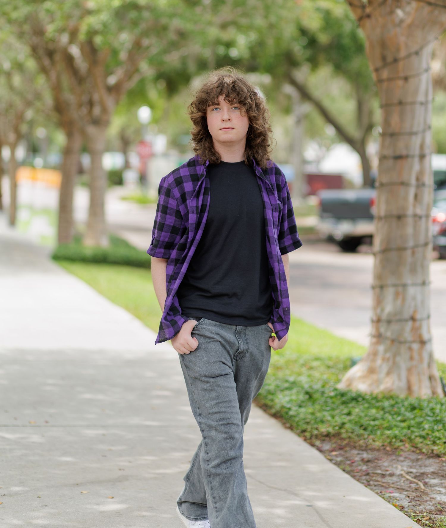 Young person with curly shoulder-length hair walks along a sunny tree-lined sidewalk wearing a purple plaid shirt, black tee and gray jeans.