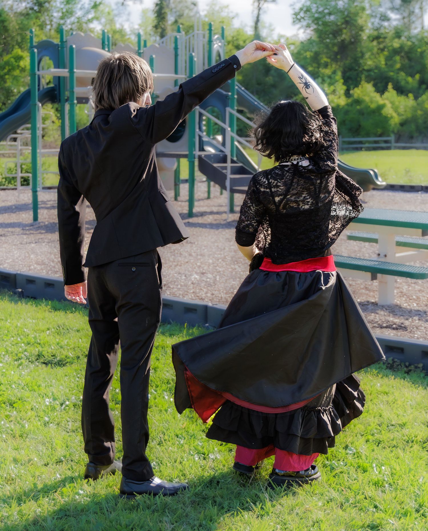 Couple dancing outdoors in black flamenco attire near a playground on a sunny day.