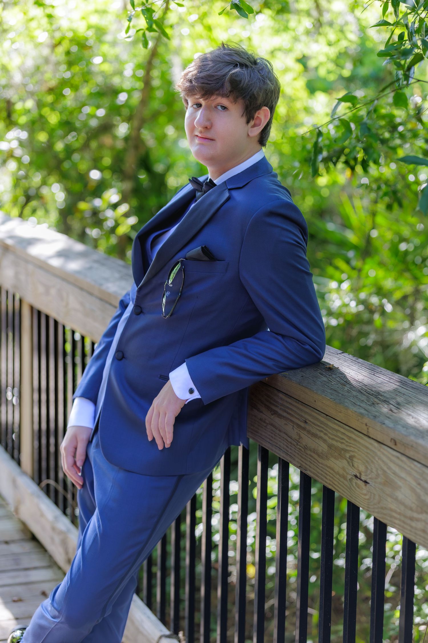 Young man in blue suit with bow tie leaning against wooden bridge railing outdoors.