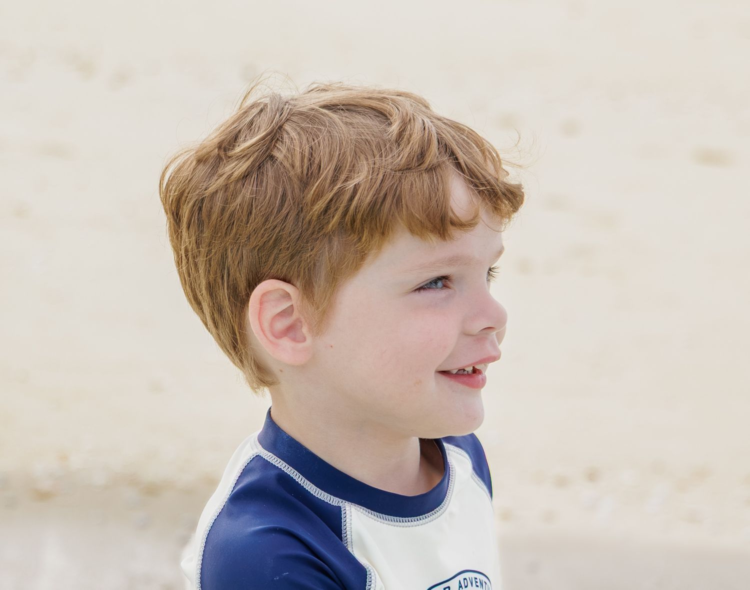 Young boy with tousled light brown hair smiling in a navy and white rash guard on a sandy beach.