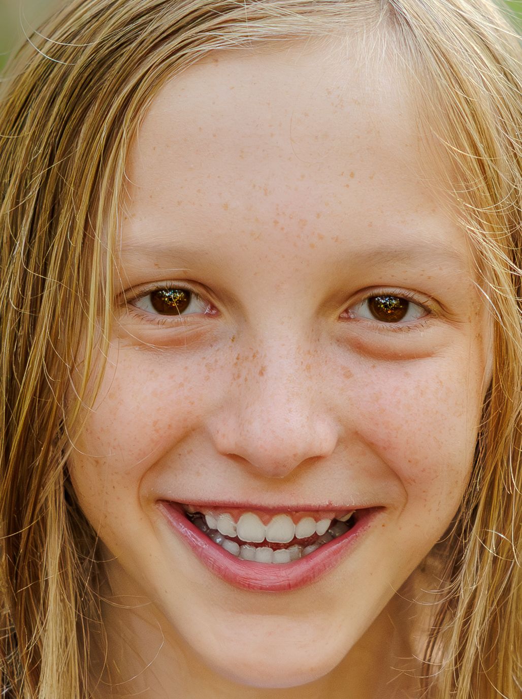 Close-up portrait of a smiling young girl with freckles, long blonde hair and brown eyes, showing a bright, cheerful expression.