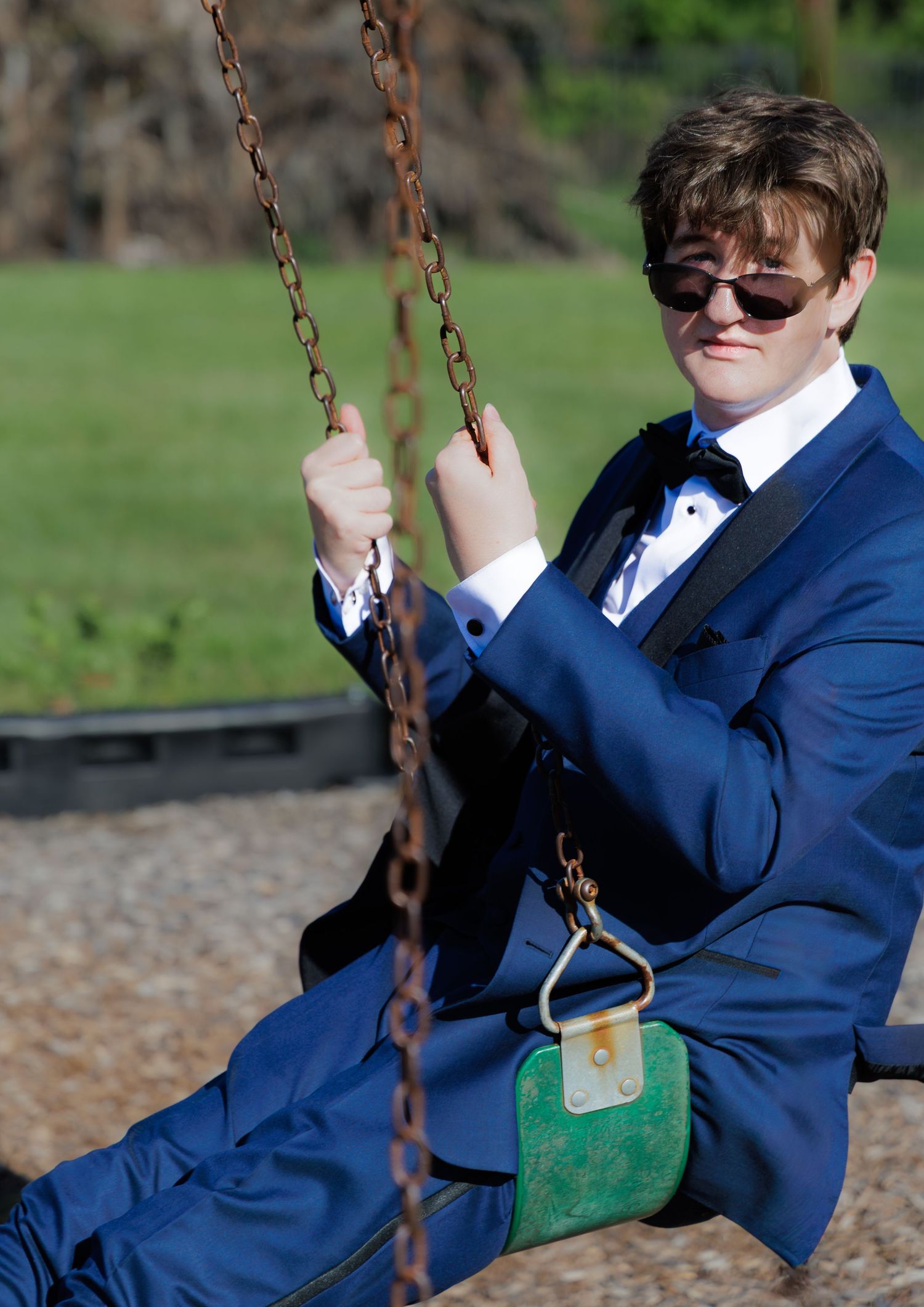 Teen in blue tuxedo and sunglasses sitting on a rusty chain swing at a playground.