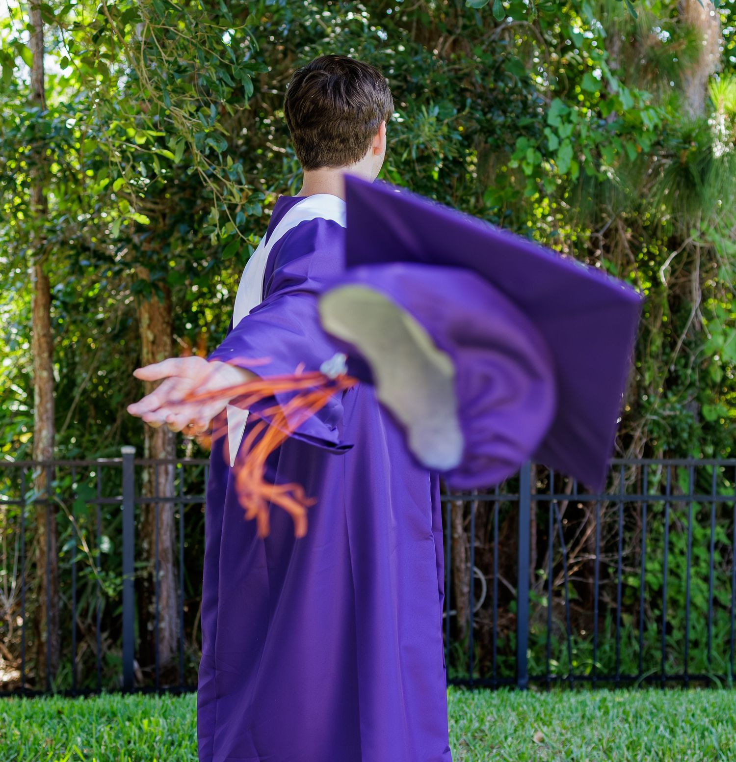 Graduate in purple gown tossing mortarboard cap into the air outdoors during graduation celebration.