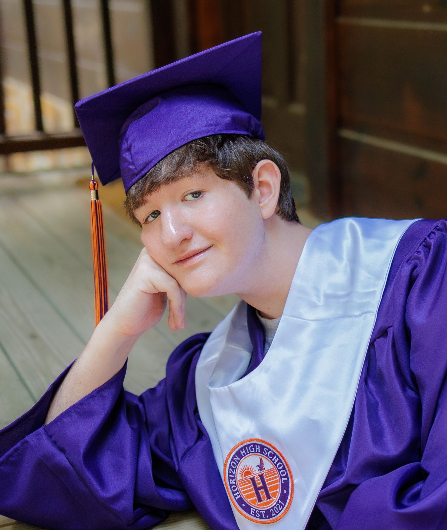 Smiling high school graduate in a purple cap and gown with a white stole resting his chin on his hand on a wooden porch.