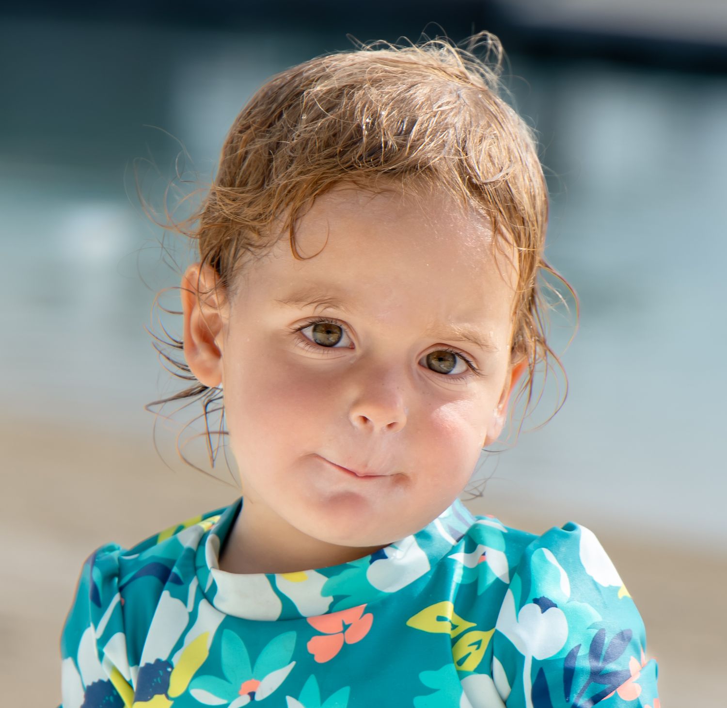 Toddler with damp reddish-brown hair wearing a teal floral swim shirt, posing on a sunny beach with soft blurred water behind.