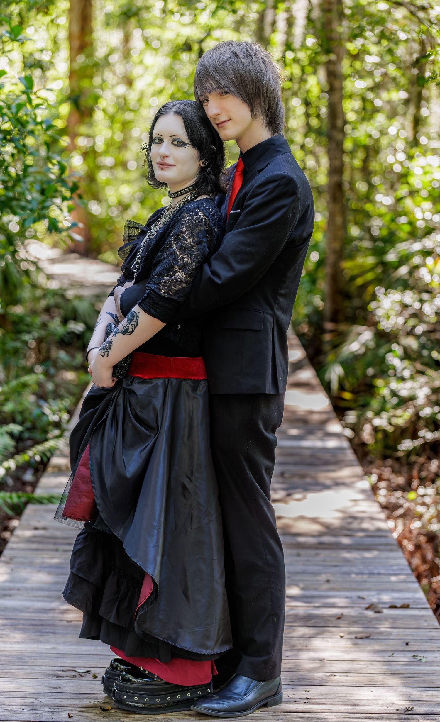 A couple in gothic formal attire poses on a woodland boardwalk, woman in black lace and man in black suit with red tie.