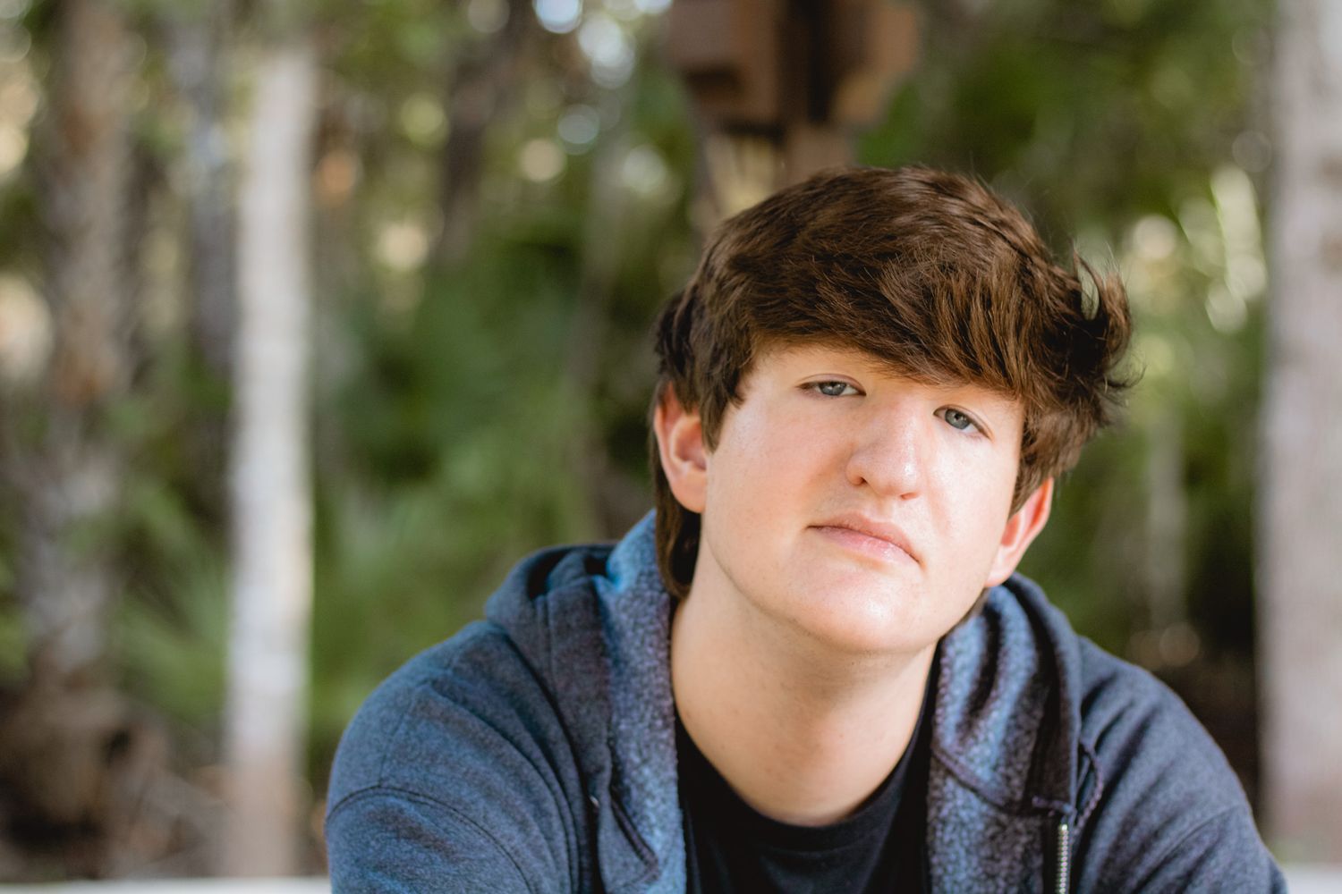 A person wearing a blue hoodie sits outdoors with trees in the background on a sunny day.