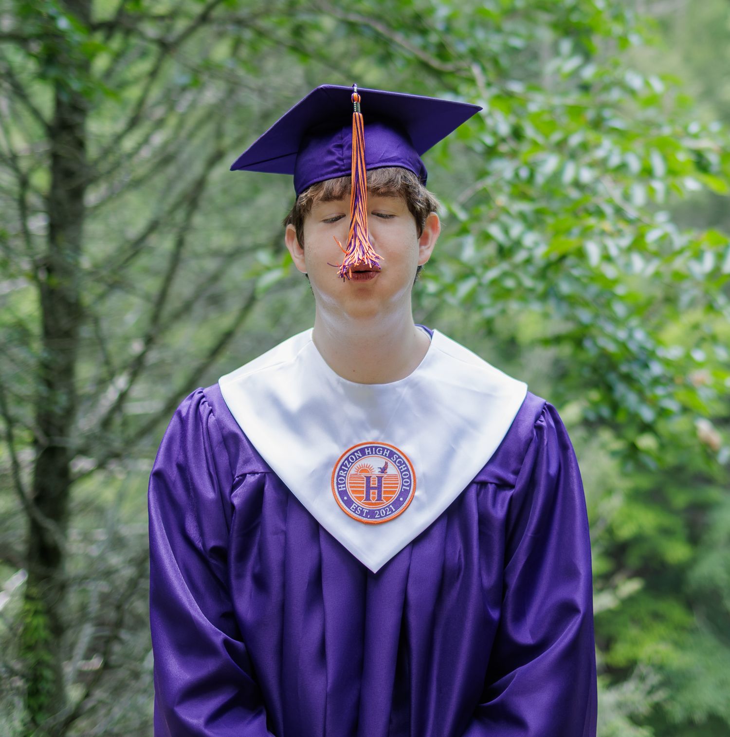 Graduate in a purple cap and gown with white stole and tassel hanging over the face, standing outdoors among green trees.