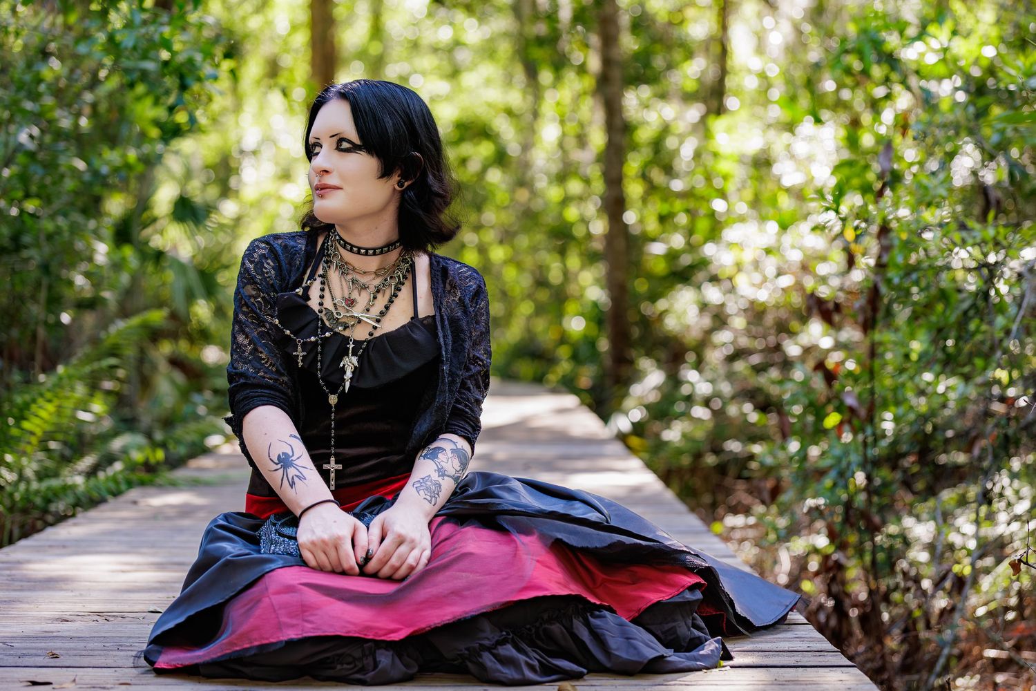 Gothic woman in black lace and red skirt sitting on wooden boardwalk surrounded by lush green forest.