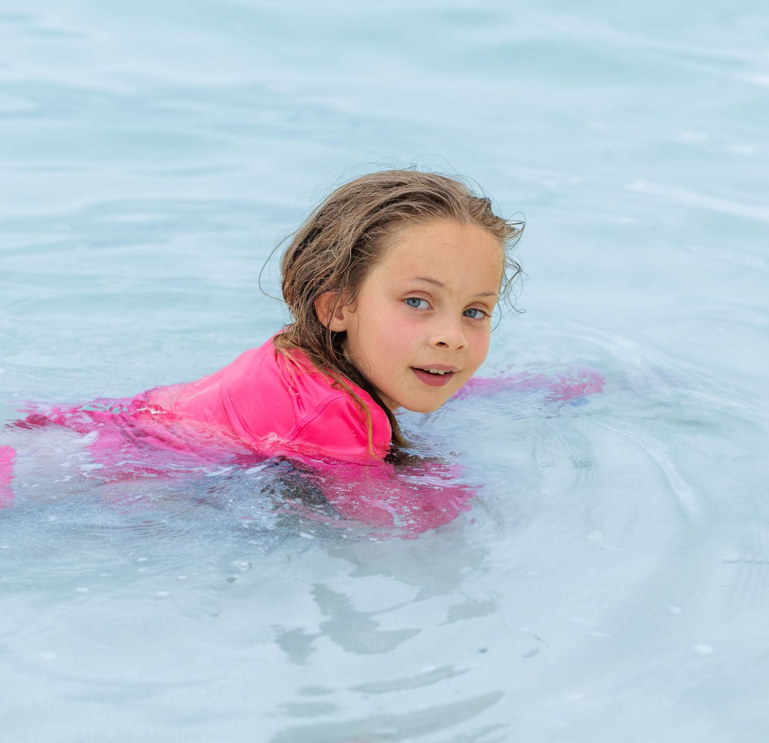Young girl in a bright pink swim shirt smiling as she swims through clear, calm blue water.