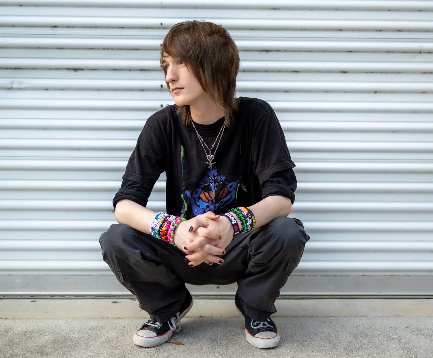 A person wearing black clothing and colorful bracelets crouches against a white corrugated metal wall.