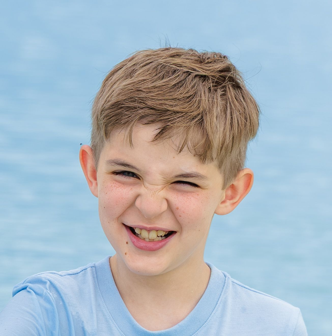 Smiling young boy with short brown hair and a squinting grin wearing a light blue t-shirt against a soft blue water background.