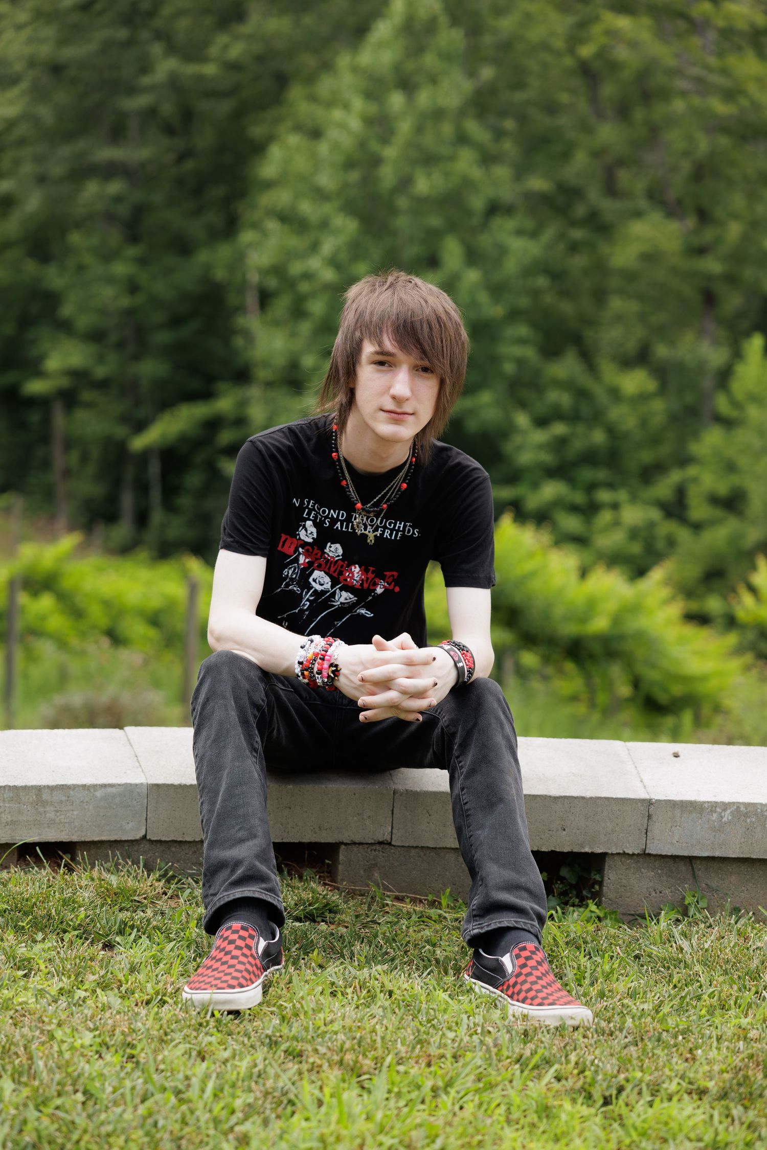 Young person with shaggy brown hair, black graphic tee, gray jeans and red checkered sneakers sitting on a stone ledge near green trees.