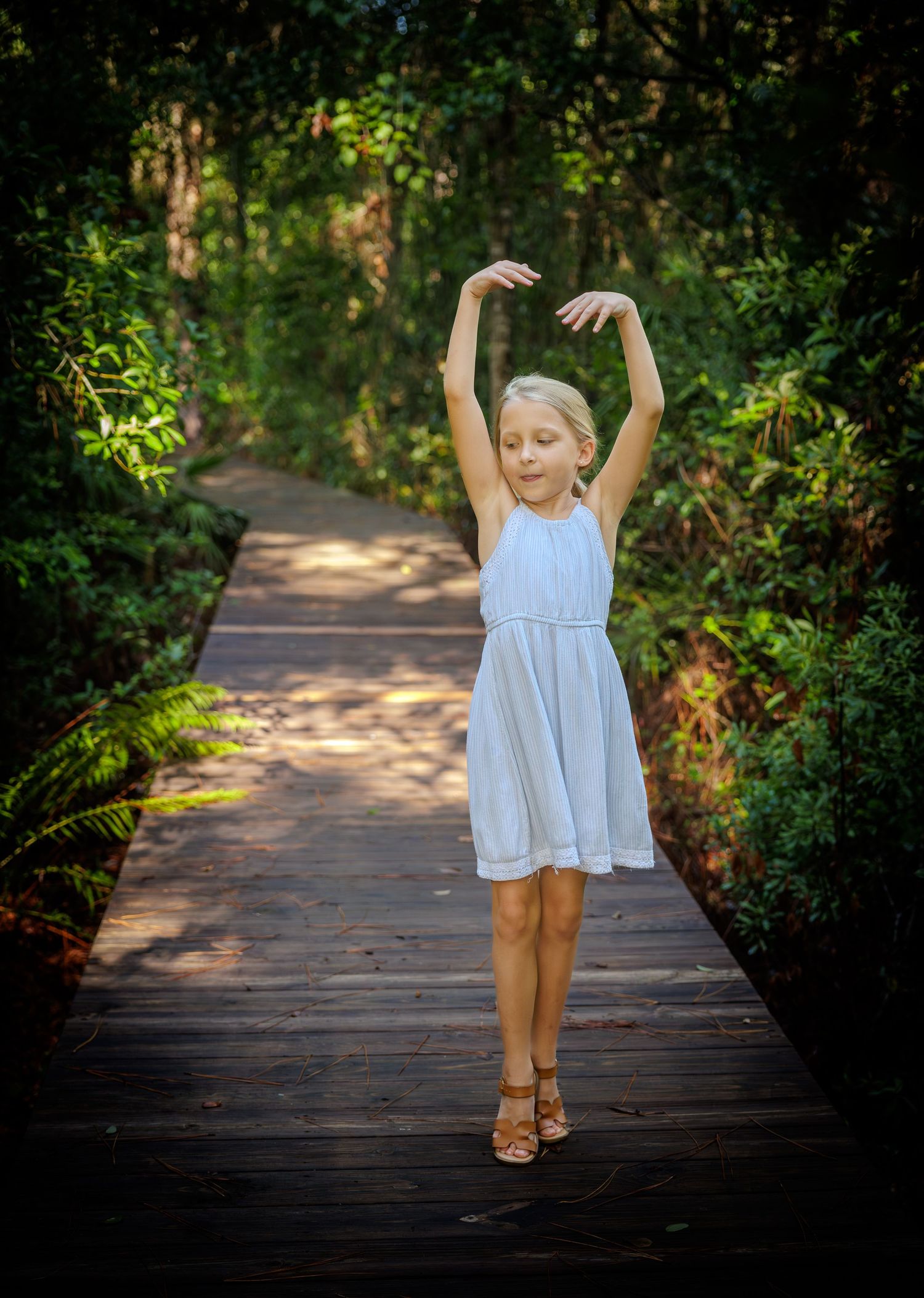 Young girl in a white dress poses in a ballet stance with arms raised on a wooden boardwalk through a sunlit green forest.