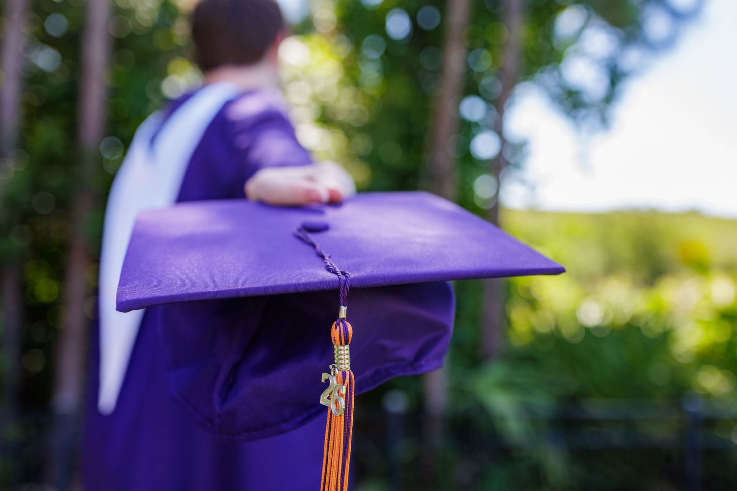 Purple graduation cap held by a graduate in gown outdoors with green trees in background.