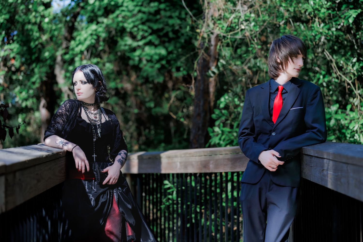 Two cosplayers on a wooden bridge, one in gothic black dress, other in dark suit with red tie, surrounded by green trees.