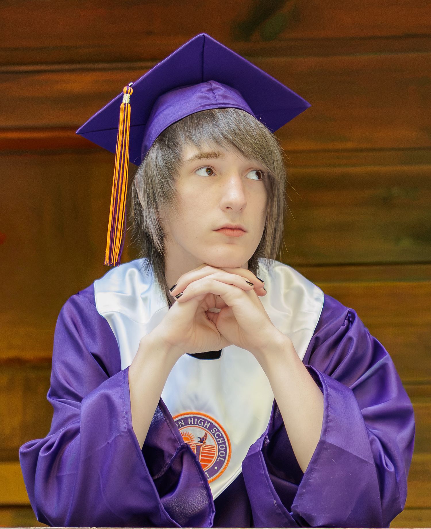 Graduate in a purple cap with orange tassel and purple gown sits with clasped hands under chin, gazing thoughtfully to the side.