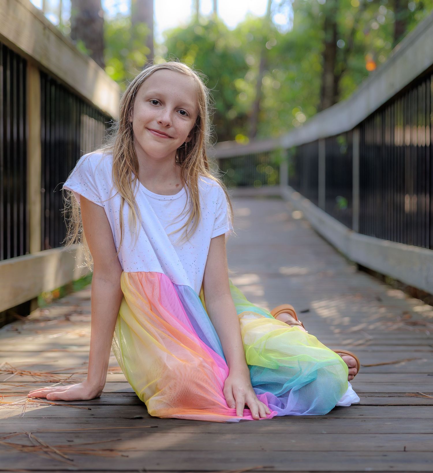Smiling girl in a rainbow pastel tie-dye dress sitting on a wooden boardwalk bridge with railings surrounded by green trees.