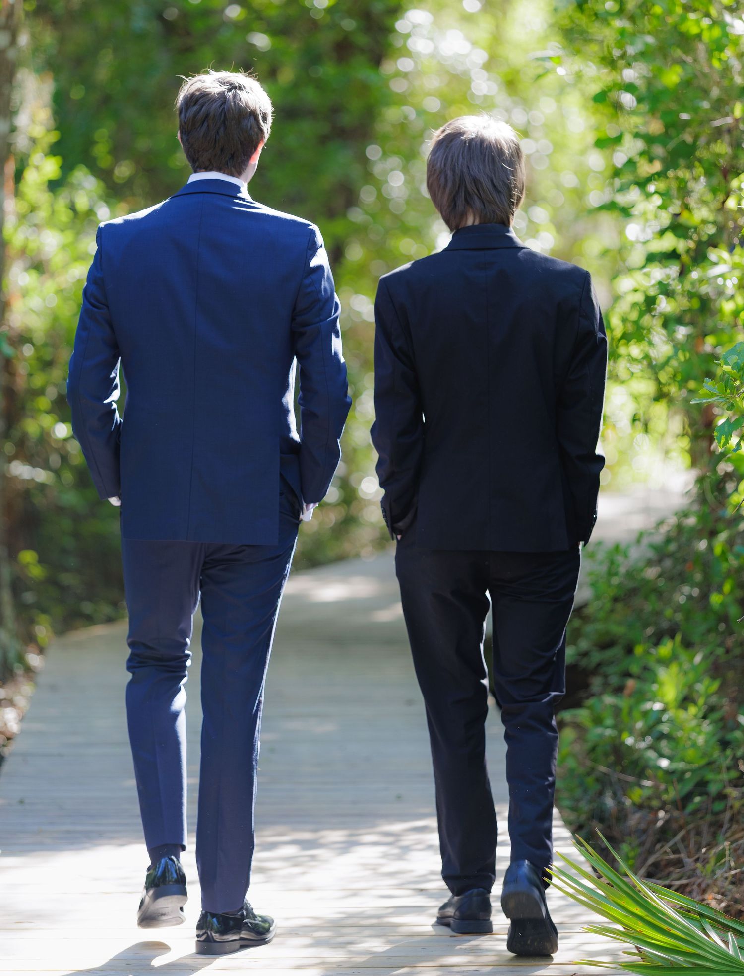 Two people in formal suits walking along a sunlit garden path surrounded by lush green trees.