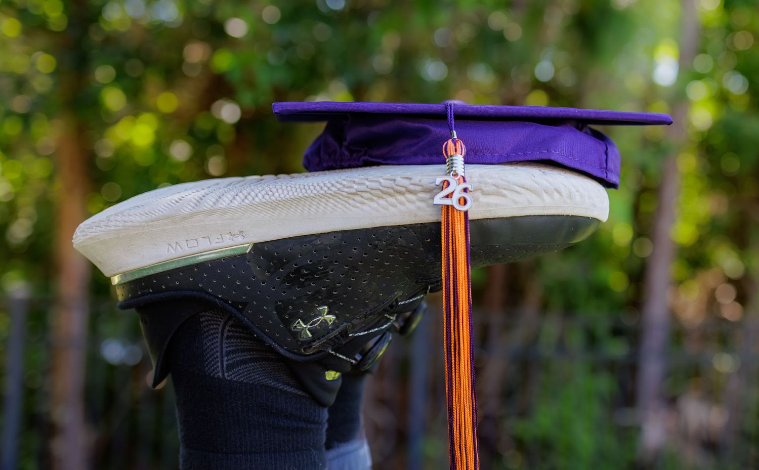 Purple graduation cap with orange tassel balanced on top of a sneaker outdoors.